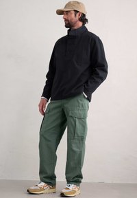 Svart skjorta med hög krage, gröna cargo-byxor med flera fickor och beige sneakers med orange accenter.
