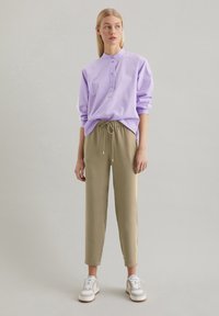 Camicia viola chiaro a maniche lunghe con colletto alto e lista di bottoni, abbinata a pantaloni khaki rilassati con cordoncino e scarpe da ginnastica bianche.