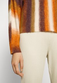 Hand naast geribde crèmekleurige broek, met een gestreepte gebreide trui met verticale strepen in oranje, bruin en wit.