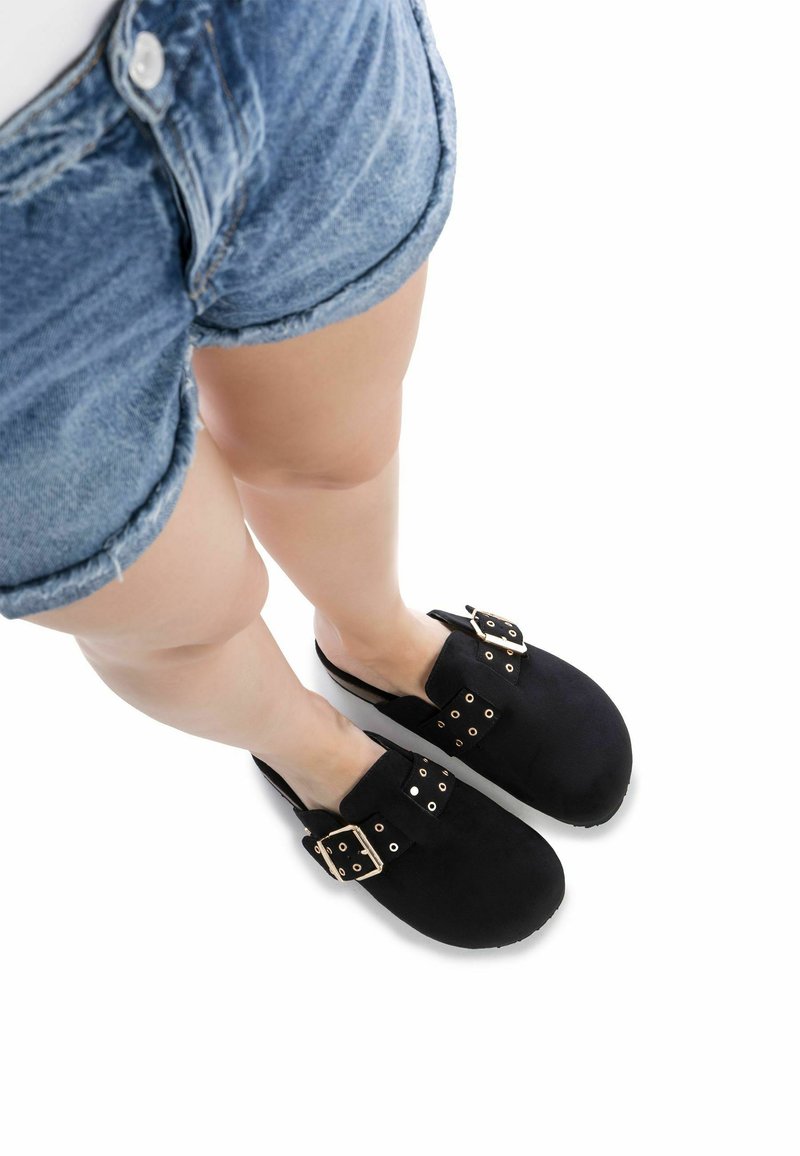 Zuecos de ante negro con hebillas y tachuelas doradas, punta redonda y diseño de parte trasera abierta, llevados con pantalones cortos de denim.