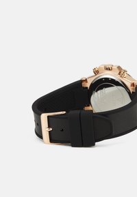 Montre chronographe avec un bracelet en cuir texturé noir, un boîtier en métal doré et un fond en acier inoxydable poli. Détail de fermoir visible.