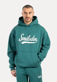 Sudadera color turquesa hecha de tejido suave, con un bolsillo frontal y un logo "Smilodox" blanco. Completa con capucha ajustable.