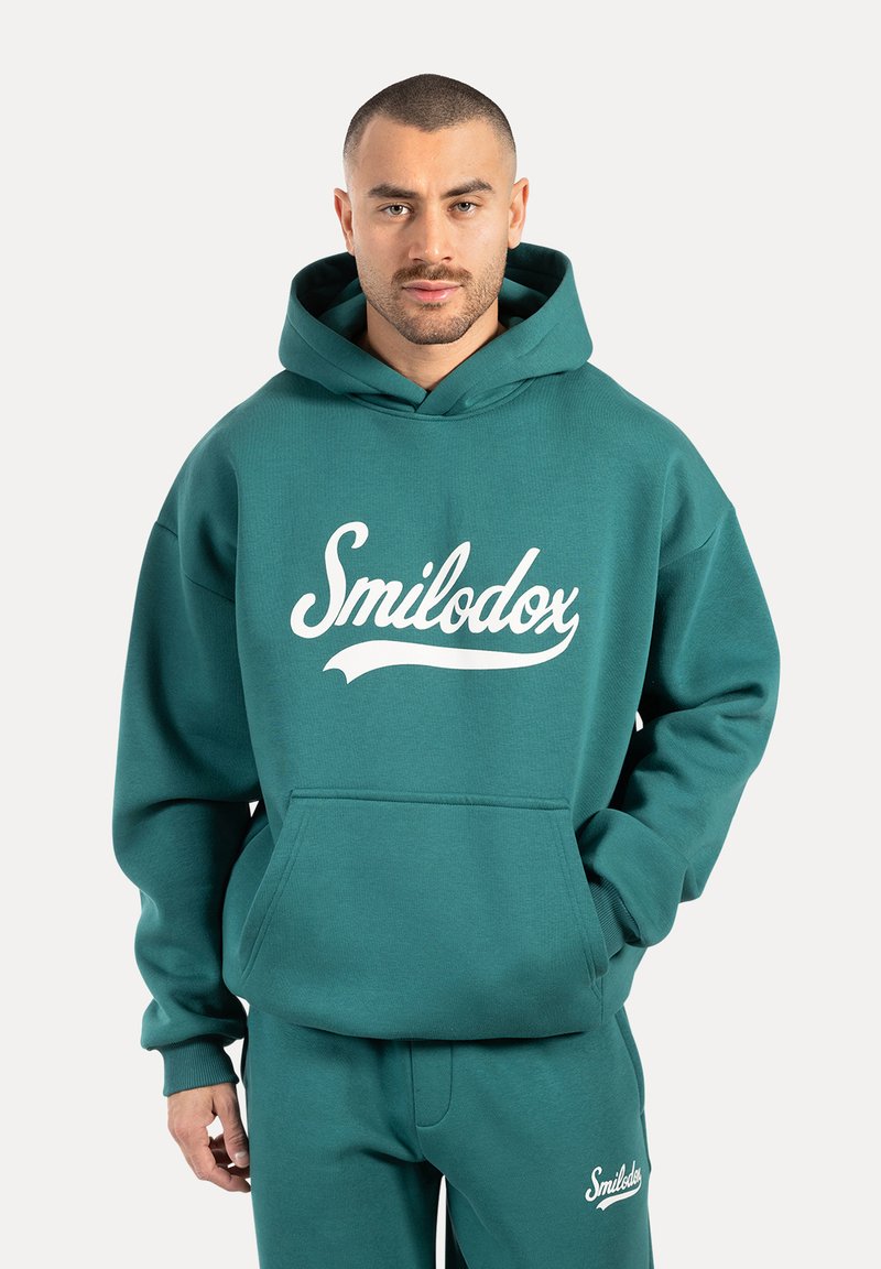 Sudadera color turquesa hecha de tejido suave, con un bolsillo frontal y un logo "Smilodox" blanco. Completa con capucha ajustable.