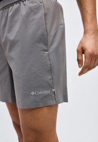 Shorts de sport Columbia gris avec poches latérales portés par une personne, montrant une ceinture élastique et un logo près de l'ourlet.
