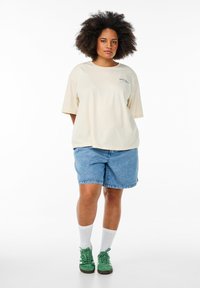 Cremefarvet oversized t-shirt med korte ærmer, parret med lyseblå denimshorts, grønne sneakers og hvide ankelstrømper. Simpelt design og pasform.