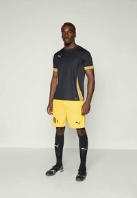 Svart og gul fotballdrakt. Trøya har en svart topp med gullakenter og et spiller nummer, kombinert med gule shortser og svarte sokker.