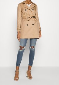 Beżowy dwurzędowy płaszcz trench z paskiem w talii, epauletami na ramionach i czarnymi guzikami, zestawiony z jasnobłękitnymi, podartymi rurkami oraz białymi szpilkami.