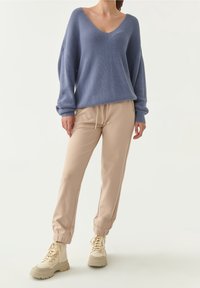 Femme portant un pull ample bleu à col en V, un pantalon de jogging beige à cordon de serrage, et des bottes à lacets épaisses couleur crème, debout sur un fond blanc.