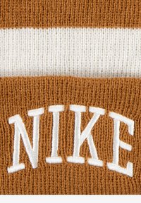 Bonnet tricoté marron et crème avec une épaisse bande blanche et un logo "NIKE" en relief. Texture douce et forme arrondie pour la chaleur.