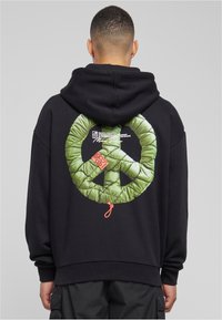 Černá oversized mikina s velkým zeleným prošívaným symbolem míru na zadní straně, s červeným štítkem umístěným blízko spodního okraje.