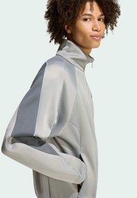 Svetlosivá bunda na zips s vysokým golierom, so modrými akcentmi pozdĺž švov a vreciek. Mäkčená tkanina s hladkou textúrou.