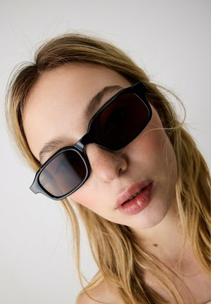 Jonge vrouw met blond haar, zwarte rechthoekige zonnebril en een neutrale uitdrukking, tegen een eenvoudige lichte achtergrond.