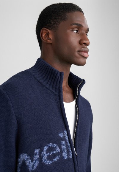 Mörkblå zip-up tröja med hög ribbad krage. Har en mjuk textur och ljusblå bokstäver över bröstet.
