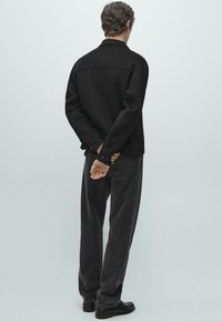 Sort uldjakke med knappedetaljer, kombineret med mørkegrå denimbukser. Modellen står med hænderne foldet bag ryggen og fremviser rygdesignet.