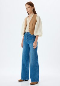 Chaleco sin mangas beige sobre una blusa blanca, combinado con jeans de mezclilla azul de pierna ancha y zapatos de tacón marrones. Diseño simple y telas naturales.