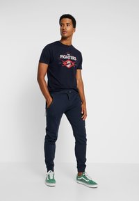 Heren navy T-shirt met een "Foo Fighters" graphic, gecombineerd met navy joggers met zijzakken en groene sneakers met witte zoolaccenten.