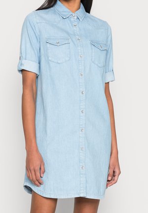 Femme portant une robe-chemise en denim bleu clair avec des manches courtes retroussées, une patte de boutonnage devant, et deux poches poitrine à rabats.