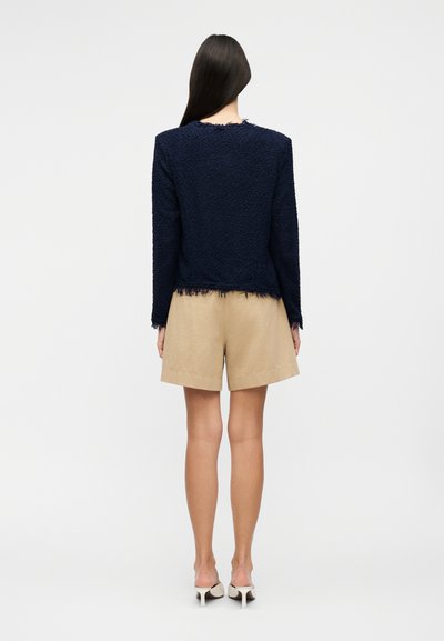 Γυναίκα που φοράει μπλε σκούρο μακρυμάνικο μπλουζάκι με υφή και ξεφτισμένα τελειώματα, συνδυασμένο με μπεζ σορτς μέχρι το γόνατο και λευκά ψηλοτάκουνα παπούτσια, στέκεται ακίνητη.