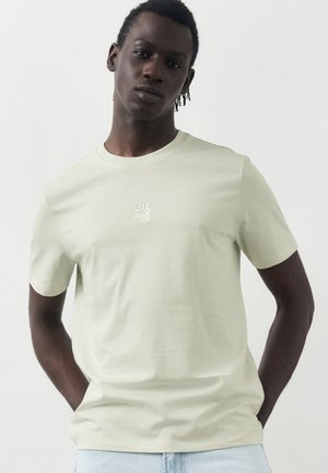 Νέος Μαύρος άνδρας με κοντά twists που φοράει ένα ανοιχτό μπεζ μπλουζάκι με μικρό λευκό κεντητό λογότυπο, στέκεται μπροστά σε λευκό φόντο.