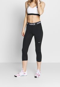 Czarne legginsy capri z szerokim, markowym pasem, zestawione z białym sportowym biustonoszem. Buty są białe i różowe z wzorzystym wzorem.