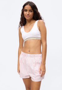 Noor naine pikkade lokkis juustega, kannab valget Calvin Kleini spordirinnahoidjat ja roosade triipudega lahtisi lühikesi pükse, seisab ühevärvilise tausta ees.