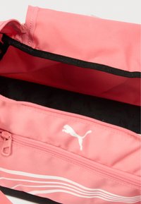 Ροζ αθλητική τσάντα με ομαλή υφή, με λευκές οριζόντιες ρίγες και λογότυπο Puma. Η εσωτερική επένδυση είναι μαύρη με επαναλαμβανόμενο μοτίβο λογοτύπου.