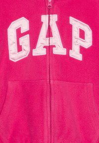 Jachetă din fleece roz cu fermoar frontal, literele "GAP" brodate în alb, centrate pe piept, și buzunare în partea de jos.
