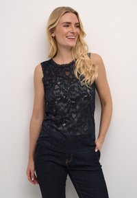 Jūras spēku bez Piedurknēm augšdaļa ar mežģīņu pārklājumu, viļņotu dekoltē un gludu tekstūru. Valkāta ar tumšiem džinsiem, kuriem ir šuvju akcenti.