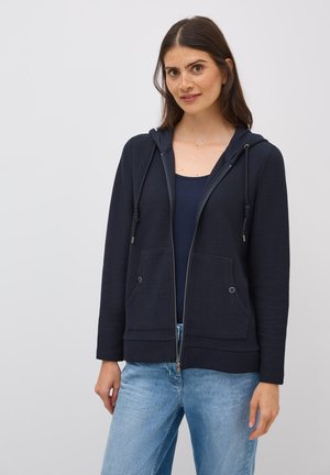Sweat à capuche zippé navy en tissu texturé, avec poches avant double, et capuche réglable à cordon, accompagné d'un jean bleu clair.