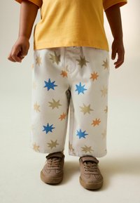 Weiße Hose mit bunten Sternmustern in Blau, Orange und Beige; kombiniert mit einem gelben Hemd und braunen Turnschuhen.