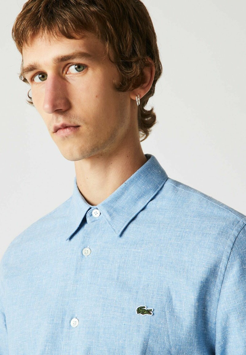 Hellblaues Poloshirt aus strukturiertem Stoff, mit einem kleinen, grünen, gestickten Logo auf der Brust und weißen Knöpfen.