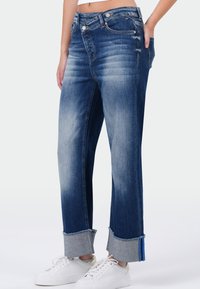 Blauwe denim jeans met een relaxte pasvorm, voorzien van een vervaagde kleur, opgerolde boorden, zichtbare zakken en metalen knoopaccenten. Gecombineerd met witte schoenen.