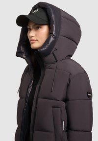 Polstret jakke i mørk lilla med høj krave og stor hætte. Funktioner inkluderer lynlås lukning, justerbare snore, og et logo patch.