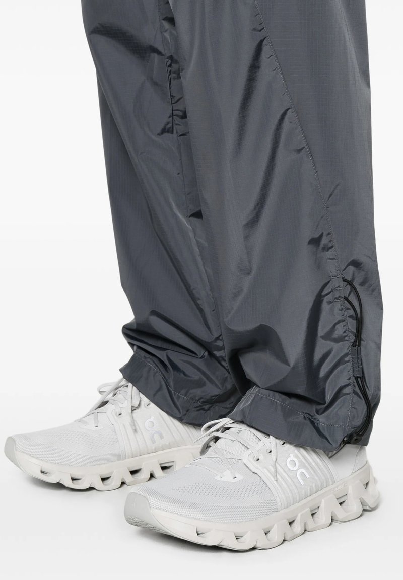 Scarpe da ginnastica bianche con suola testurizzata indossate con pantaloni grigio scuro regolabili con coulisse.