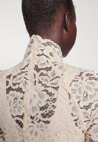 Lace blouse in beige featuring floral patterns, high neckline, and a button closure at the back. Textured, intricate detailing throughout.