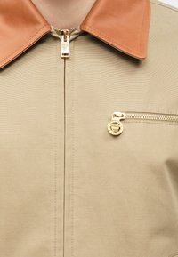 Veste beige avec col en cuir marron clair et détail de fermeture éclair dorée au centre ainsi qu'une petite poche poitrine zippée avec une tirette circulaire.