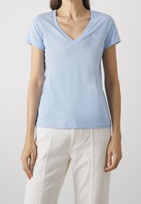 Ljusblå bomullströja med v-ringad hals, korta ärmar och en liten broderad logotyp på bröstet. Bärs med vita byxor.