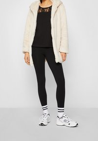 Person som bär en vit fluffig jacka, svart linne med spets, svarta leggings, vita strumpor med svarta ränder och vita New Balance-skor.