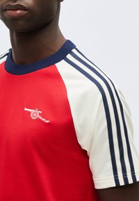 Άνδρας με κόκκινο και λευκό κοντομάνικο πουκάμισο με σκούρο μπλε στρογγυλή λαιμόκοψη και ρίγες στους ώμους, με μικρό έμβλημα κανονιού στο στήθος.