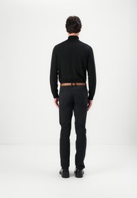 Schwarzes Langarmoberteil mit hohem Kragen, kombiniert mit dunklen Hosen. Ein brauner Ledergürtel betont die Taille. Schwarze Schuhe runden das Outfit ab.