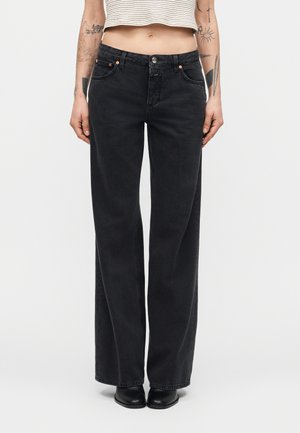 Person iført højtaljede, vide sorte jeans, kortstribet top og sorte sko, med synlige armtatoveringer mod en ensfarvet baggrund.