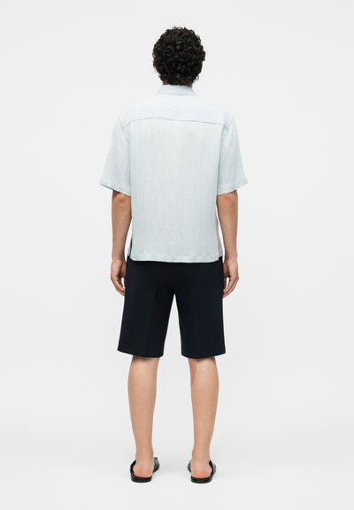 Άνδρας με σγουρά μαλλιά που φοράει ένα ανοιχτό μπλε κοντομάνικο πουκάμισο, μαύρα σορτς μέχρι το γόνατο και μαύρα σανδάλια slide, με την πλάτη γυρισμένη σε λευκό φόντο.