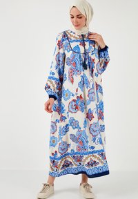 Rochie lungă, florală, în nuanțe de crem, albastru și roșu, cu un guler înalt, mâneci bufante și accente de ciucuri. Purtați cu adidași într-o nuanță neutră.