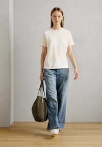 T-shirt a maniche corte crema, vestibilità rilassata, abbinato a jeans a gamba larga celesti. Tiene una borsa grigia, in piedi su un pavimento in legno contro una parete neutra.