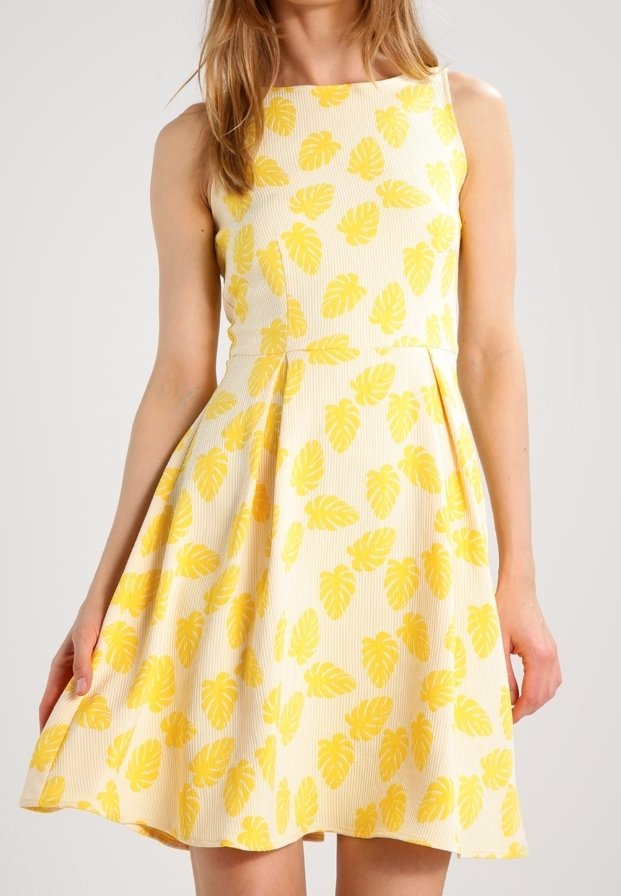Femme portant une robe blanche sans manches, longueur genou, avec un motif de feuilles tropicales jaunes, debout devant un fond clair uni.