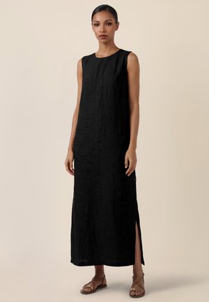 Frau trägt ein ärmelloses schwarzes Maxikleid mit Seitenschlitz und braune Riemchensandalen, steht vor einem schlichten beigen Hintergrund.