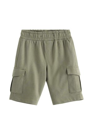 Drenge står smilende iført olivengrøn T-shirt med legende frugtmønster og matchende shorts, beige slip-on sko, ensartet lyst baggrund.