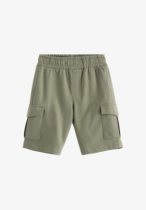 Drenge står smilende iført olivengrøn T-shirt med legende frugtmønster og matchende shorts, beige slip-on sko, ensartet lyst baggrund.