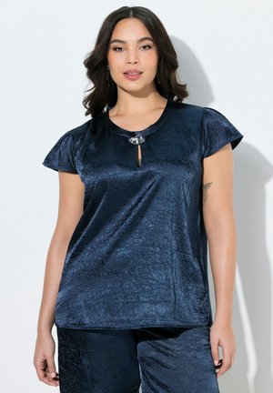 Nainen, jolla on tummat, laineikkaat hiukset, pukeutunut kiiltävään tummansiniseen lyhythihaiseen toppiin ja sitä vastaaviin housuihin, seisoo valkoista taustaa vasten.