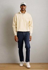 Mann trägt einen hellbeigen Halb-Zip-Sweatshirt, dunkelblaue Hose, weiße Sneakers und eine weiße Kappe, während er vor einer einfachen hellen Wand und einem Holzboden steht.